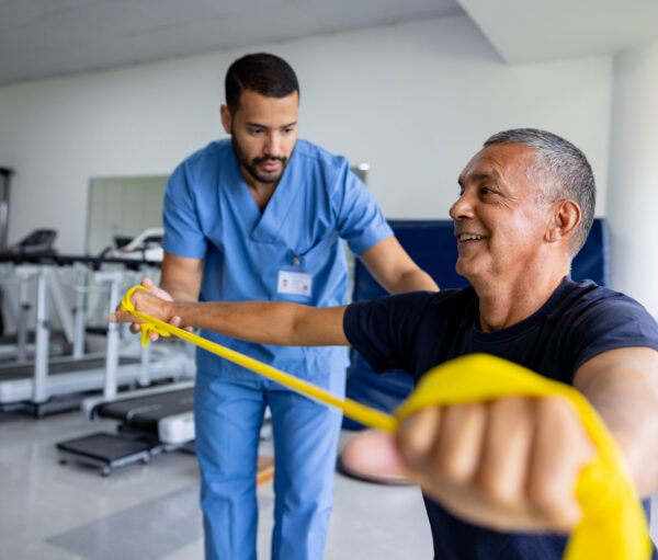 Mature Latin American man doing physical therapy exercises using a stretch band with the assistance of his therapist